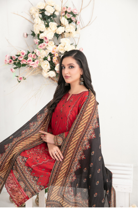Woman wearing a red outfit with a black and gold patterned dupatta, standing against a white wall with floral decorations.