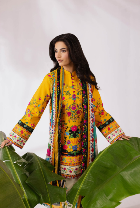 Woman wearing a yellow embroidered traditional outfit with a colorful scarf, standing behind large green leaves.