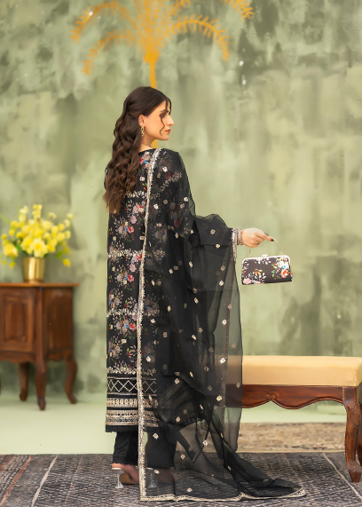 Woman in a black traditional outfit with floral patterns, holding a clutch, against a decorative indoor background.