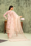 Woman in a light pink traditional outfit with a sheer dupatta, holding a floral bag against a textured green wall.