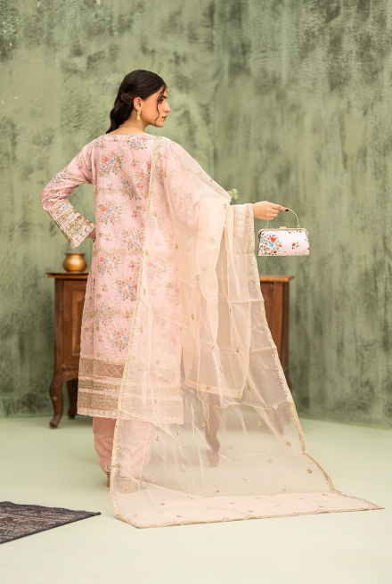 Woman in a light pink traditional outfit with a sheer dupatta, holding a floral bag against a textured green wall.