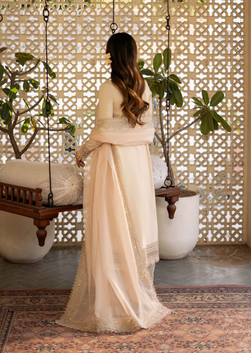 Woman in a beige saree standing in a decorated room with plants and a lattice wall.