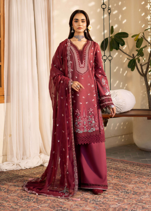 Woman in a burgundy embroidered traditional outfit standing in a well-lit room with white curtains and a plant.