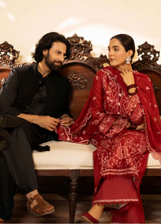 Man and woman sitting on an ornate wooden bench, man wearing black and woman in red traditional outfit.