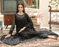 Woman in a black traditional outfit sitting on a patterned rug with a vintage television and decorative items in the background.