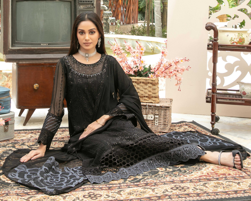 Woman in a black traditional outfit sitting on a patterned rug with a vintage television and decorative items in the background.