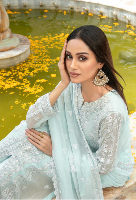 Woman in a light blue embroidered outfit by a fountain with yellow flowers.