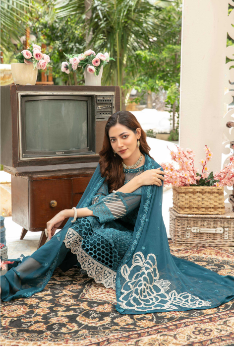 Woman in a teal traditional outfit sitting on a patterned rug with a vintage television and flowers in the background.