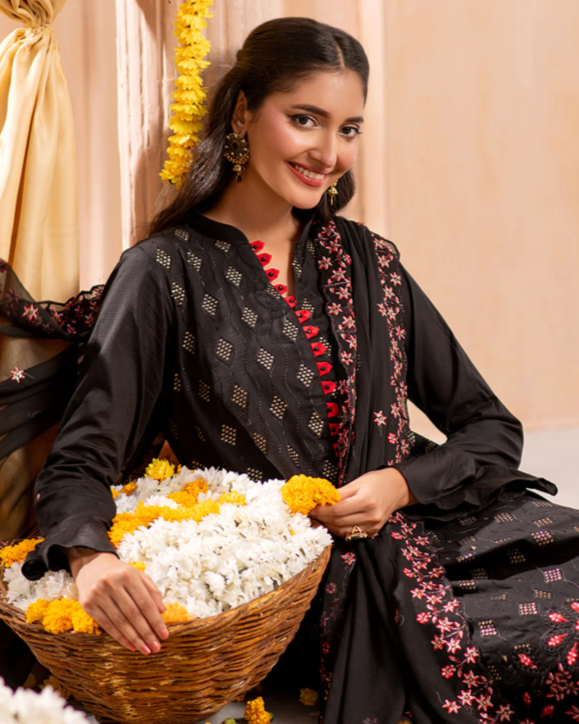 Woman in a black embroidered outfit holding a basket of flowers against a beige curtain.