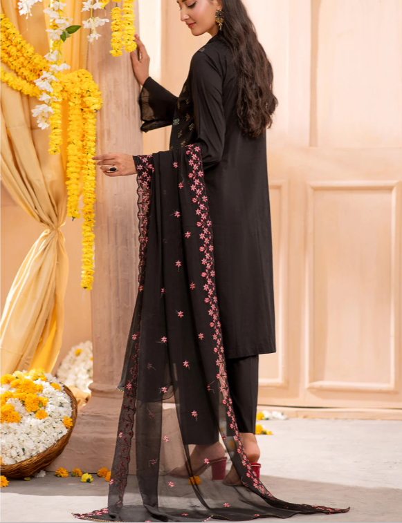 Woman in a black traditional outfit with floral decorations, standing against a beige wall.